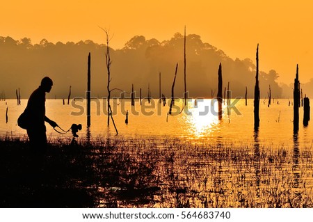 A photographer standing by the lake over viewing the dead and sunken tree in the water taking photos during sunset located at Kenyir Lake, Terengganu, Malaysia. ( grain and soft effect)