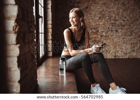 Picture of attractive fitness woman sitting in gym while using phone and listening music with earphones.