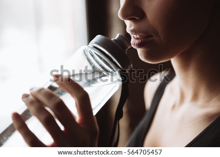 Cropped picture of gorgeous young fitness woman standing in gym near window while drinking water.