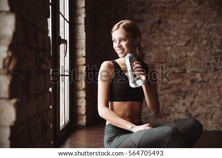 Photo of happy fitness woman sitting in gym while drinking water and look aside.