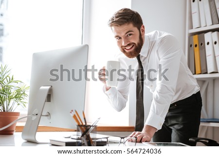 Picture of attractive young businessman dressed in white shirt standing in office while drinking tea and using computer.