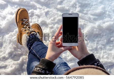 Close up of a young beautiful hipster girl using white mobile smart phone in trendy orange boots and ripped jeans outdoor on the snow background in a cold winter day Royalty-Free Stock Photo #564792085