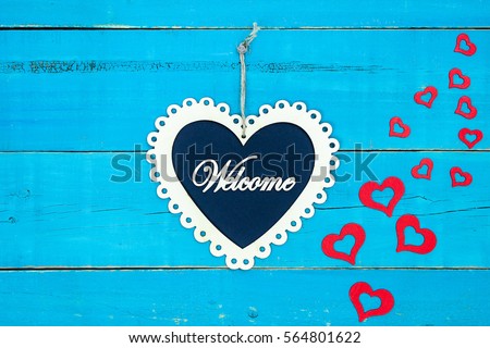 Welcome slate and white wood lace heart sign hanging from rope with cascading red hearts on rustic antique teal blue wooden background; Valentine's Day and love concept with painted copy space