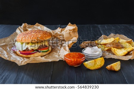 Fast food restaurant dish top view. Meat cheese burger in craft paper, potato chips and wedges. Take away set on dark black wood background. Hamburger and spicy tomato sauce.