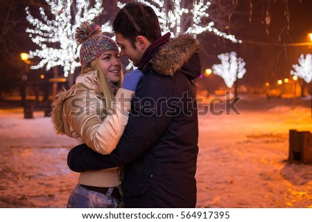 Closeup front view image of a young cheerful couple in winter clothing embrace at night during winter holidays. Luminous decoration background