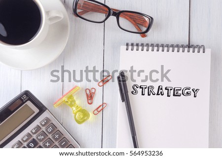 STRATEGY text on notebook.coffee,calculator,pen,rubber stamp,glasses on the desk.top view.