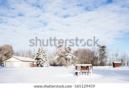 A "For Sale" sign nailed to a wooden pallet on an empty snow covered lot beside a two car garage and a boat with tall trees in the background in a winter landscape