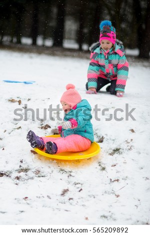 Two sisters in bright ski suits ride the sledge in winter day. Elder sister has pushed the sledge with younger sister. It is snowing. The baby is excited.