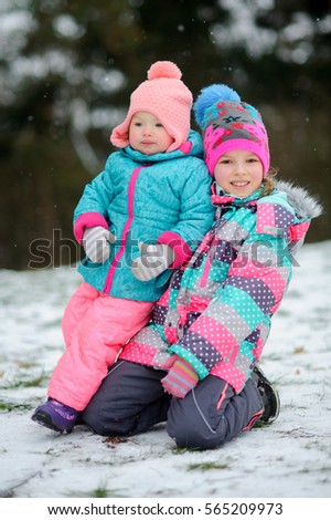 Two little girls on winter walk. The older girl stands on snow on a lap and embraces the younger girl. Children warmly are also brightly dressed. It is snowing.