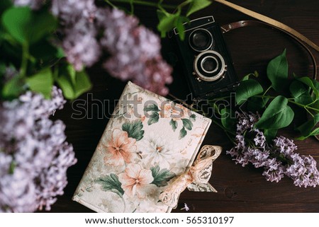 Top view of note book, lilac and old camera on dark wooden table.