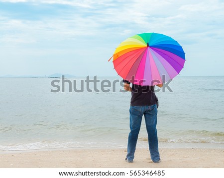 Man holding rainbow color of umbrella and looking to the sea, Asian young man holding rainbow umbrella on the beach, man walking along the bea