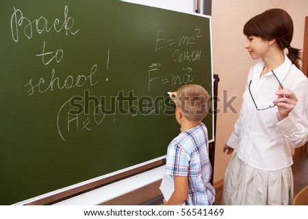 Photo of elementary student writing formulae on blackboard with his teacher near by