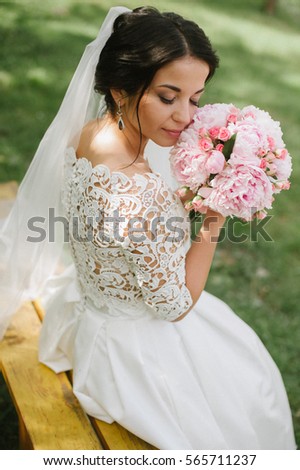 The bride sits on a bench and sniffs bouquet of peonies. Very gentle picture. Young brunette girl in wedding day. Green lawn