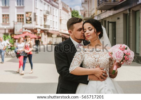 Groom kisses bride from behind standing on tram ways somewhere in old city. Beautiful romantic newlywed couple hugging near flower bouquets at luxury french restaurant outdoors.Wed  bouquet of peonies