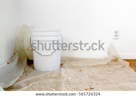 A white bucket over a sheet of plastic placed over carpet floor to collect water from leaking ceiling Royalty-Free Stock Photo #565726324