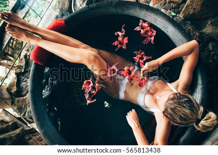 Woman relaxing in round outdoor bath with tropical flowers. Organic skin care in kawa hot bath in luxury spa resort. Royalty-Free Stock Photo #565813438