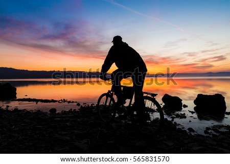Mountain bike (mtb) and biker. Silhouette of a mountain bike and of a cyclist who is watching the sunrise over the lake (lake Varese - Varese, Italy) - Focus on mountain bike and biker Royalty-Free Stock Photo #565831570