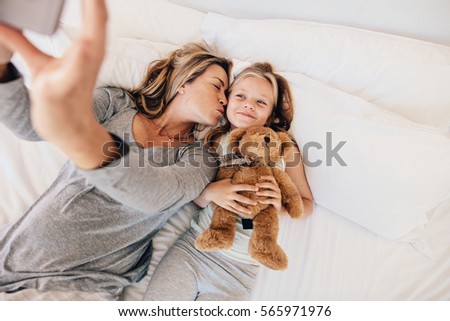 Mother and daughter lying on bed and taking self portrait with mobile phone. Woman kissing her daughter and taking self portrait.