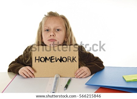 young beautiful little schoolgirl sad and overwhelmed holding paper with the text homework written sitting at school desk in stress isolated on white background in education work concept
