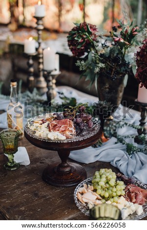 Plate of cheese with jamon and grape served with vine, standing on old wooden table decorated by bouquets of hydrangea, candles and grey cloth. Banquet. Decoration.