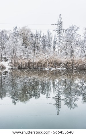 Reflection of trees covered snow in the water. Trees on the pond. The river in the winter season. Fairytale landscape. Electric high voltage tower in the trees.