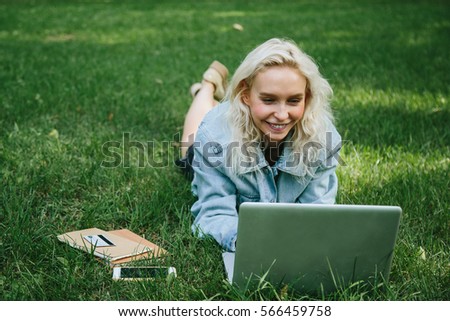 Beautiful young blonde woman lying on the green grass in park with her laptop and smiling. Student girl looking at the computer screen.  