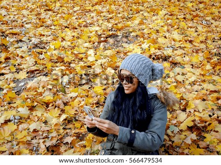 Attractive female person in winter clothes wearing sunglasses and using smartphone on autumn leaves background 
