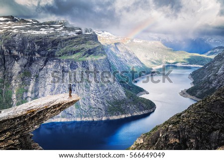 Trolltunga (Troll Tongue), climber standing at edge of cliff Trolltunga looking at rainbow against mountains, over amazing blue lake. Beautiful landscape of wild nature in Norway, Scandinavia. Royalty-Free Stock Photo #566649049