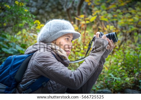 asian backpacker with camera in hands taking outdoor photo in a park with winter jacket and winter hat on 
