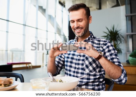 Smiling man photographing his meal in a cafeteria on his smartphone, copy space to the side over a large glass wall