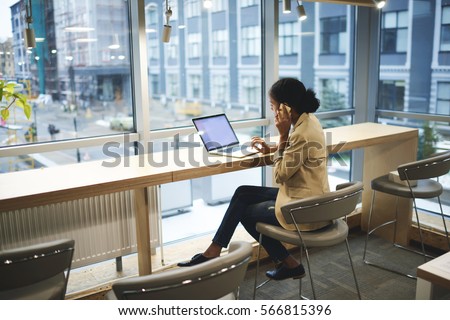 Young afroamerican executive making remote job being in business trip sitting at table in coworking space talking on the phone with entrepreneur using laptop computer with mock up screen with wifi