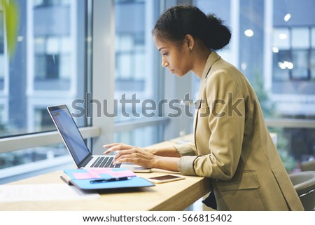 Concentrated female administrative typing messages to employee coordinating work being in business trip using laptop computer and wireless connection to internet sitting in modern coworking space