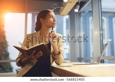 Portrait of skillful afro american female executive thinking over working schedule for employee writing report into notebook while using laptop computer and wireless connection in coworking space  Royalty-Free Stock Photo #566815405