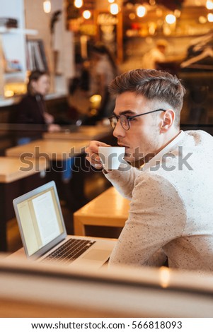Picture of attractive young man sitting in cafe and chatting by laptop computer. Looking aside while drinking coffee.