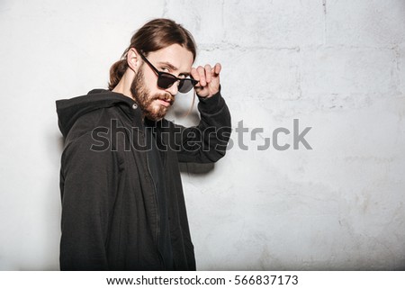 Photo of young handsome bearded hipster man wearing sunglasses posing over wall background.