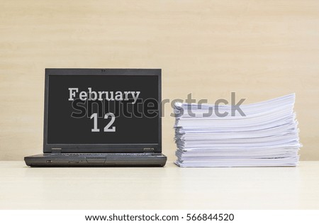 Closeup computer laptop with february 12 word on the center of screen in calendar concept and pile of work paper on wood desk and wood wall in work room textured background with copy space