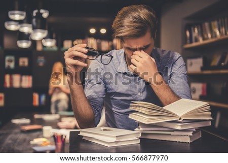 Tired young man is holding eyeglasses and massaging his nose bridge while working hard in the modern library Royalty-Free Stock Photo #566877970