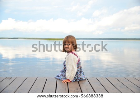 Little girl in the traditional russian dress (sarafan)  alone goes alonge  a wooden pier. View of the beautiful calm lake and clouds reflexion. 