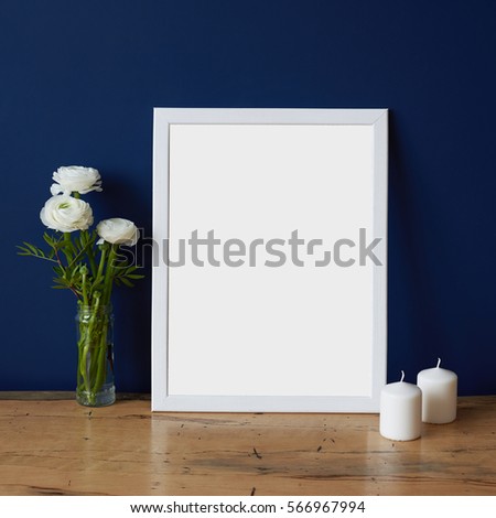 Empty modern style frame, bouquet of white flowers ranunculus and two white candles on a deep blue wall background and wooden table. Poster mockup in hipster romantic style interior.