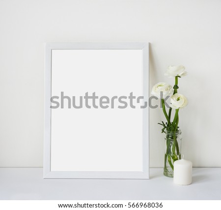 Empty white frame with place for text on the white wall and table, bouquet of flowers ranunculus and candle. Scandinavian style room interior. Template mock up for paintings or photographs.