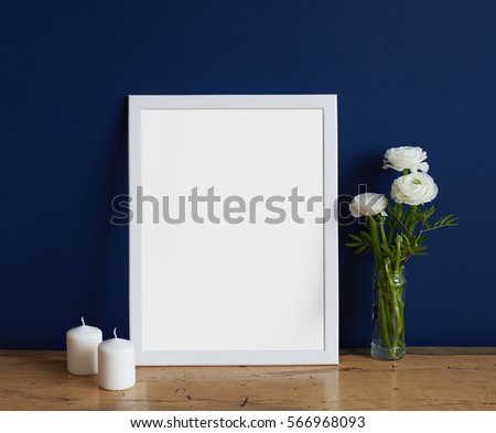Empty frame mockup for design presentation, bouquet of flowers ranunculus and two white candles on a deep blue wall background and wooden table. Hipster romantic style concept, minimalism design.
