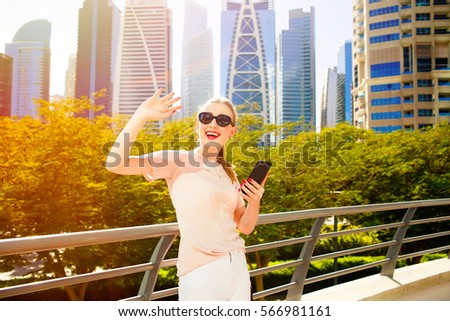 Cheerful woman with red lips raises her hand up while she stands on bridge before skyscrapers