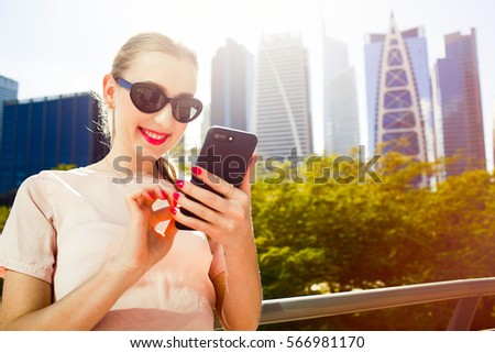 Smiling lady with pink lips checks her iPhone standing before skyscrapers in park