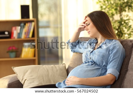 Pregnant woman with hand on forehead suffering headache sitting on a couch in the living room at home Royalty-Free Stock Photo #567024502