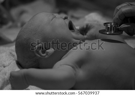Newborn baby being examined by paediatric doctors moments after birth with stethoscope and gloves checking vital signs in neonatal care crying caucasian female daughter in black and white
