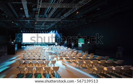 Super conference hall with white chairs and colored illumination and blank screen Royalty-Free Stock Photo #567139513