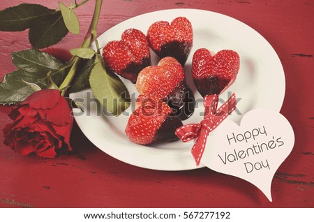 Happy Valentines Day chocolate dipped heart shaped strawberries on white plate on red vintage wood background, with red rose and gift tag, with applied retro style filters.