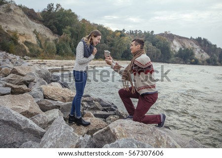 Happy thoughtful couple standing on a rock beach near sea hugging each other in cold foggy cloudy autumn weather. Man make proposal to woman with a huge rock. Copy space