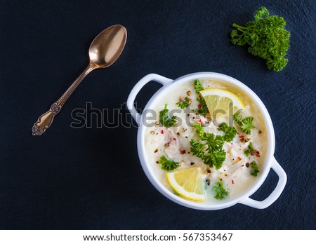 Avgolemono, chicken soup with egg-lemon sauce, rice and fresh parsley leaves in white bowl on black stone background. Top view.