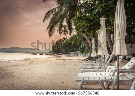 Deserted tropical beach with sunbeds and umbrellas among palm trees without people during beautiful orange sunset in hidden paradise in Thailand